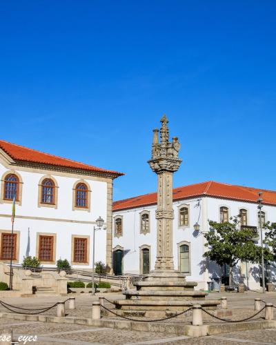 The image shows a sunlit town square in Portugal featuring a prominent stone pillory (pelourinho) in the center, flanked by white buildings with red-tiled roofs, including a municipal building with a Portuguese flag and a blue ribbon; the photo is