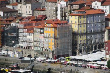 A view of the Ribeira district in Porto, Portugal, showing colorful traditional buildings along the Douro riverfront, a stone quay with market stalls under white umbrellas, and a tour boat on the water.