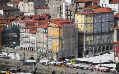 A view of the Ribeira district in Porto, Portugal, showing colorful traditional buildings along the Douro riverfront, a stone quay with market stalls under white umbrellas, and a tour boat on the water.