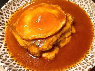 A close-up shot of a Francesinha sandwich served in a patterned ceramic bowl, featuring layers of meat and bread smothered in a thick, reddish-brown tomato and beer sauce with a fried egg on top.
