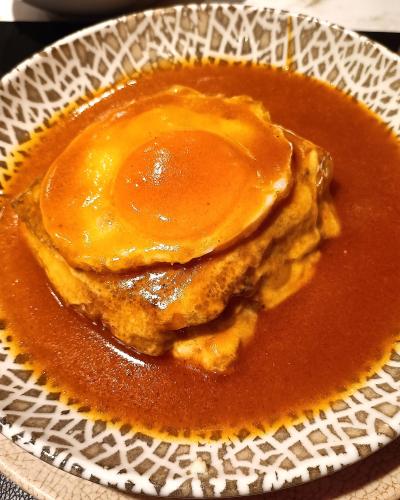 A close-up shot of a Francesinha sandwich served in a patterned ceramic bowl, featuring layers of meat and bread smothered in a thick, reddish-brown tomato and beer sauce with a fried egg on top.