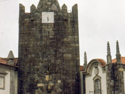 A tall, medieval stone defensive gatehouse featuring a clock face, a bell on a metal spire, and an arched passage at the base, flanked by white buildings and vintage cars.