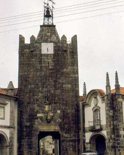 A tall, medieval stone defensive gatehouse featuring a clock face, a bell on a metal spire, and an arched passage at the base, flanked by white buildings and vintage cars.
