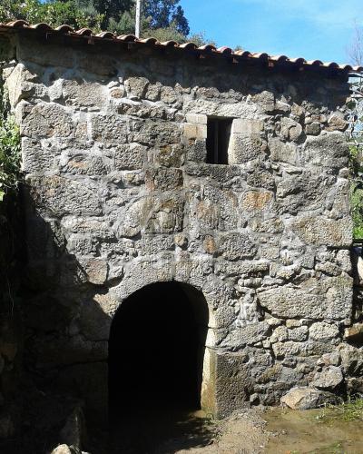 A stone water mill with a tiled roof and an arched entrance built into a stream bank, with a modern building visible in the background.