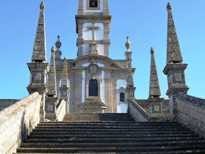 A low-angle photograph of a white church facade with a central bell tower topped by a metal cross, accessed by a long, wide stone staircase flanked by stone balustrades and four prominent obelisks.