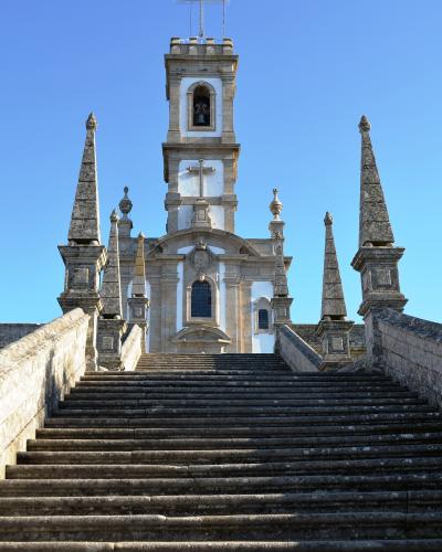 A low-angle photograph of a white church facade with a central bell tower topped by a metal cross, accessed by a long, wide stone staircase flanked by stone balustrades and four prominent obelisks.