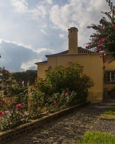 A yellow-walled building with a tiled roof and chimney, surrounded by a garden with palm trees, flowering bushes, and a cobblestone path.