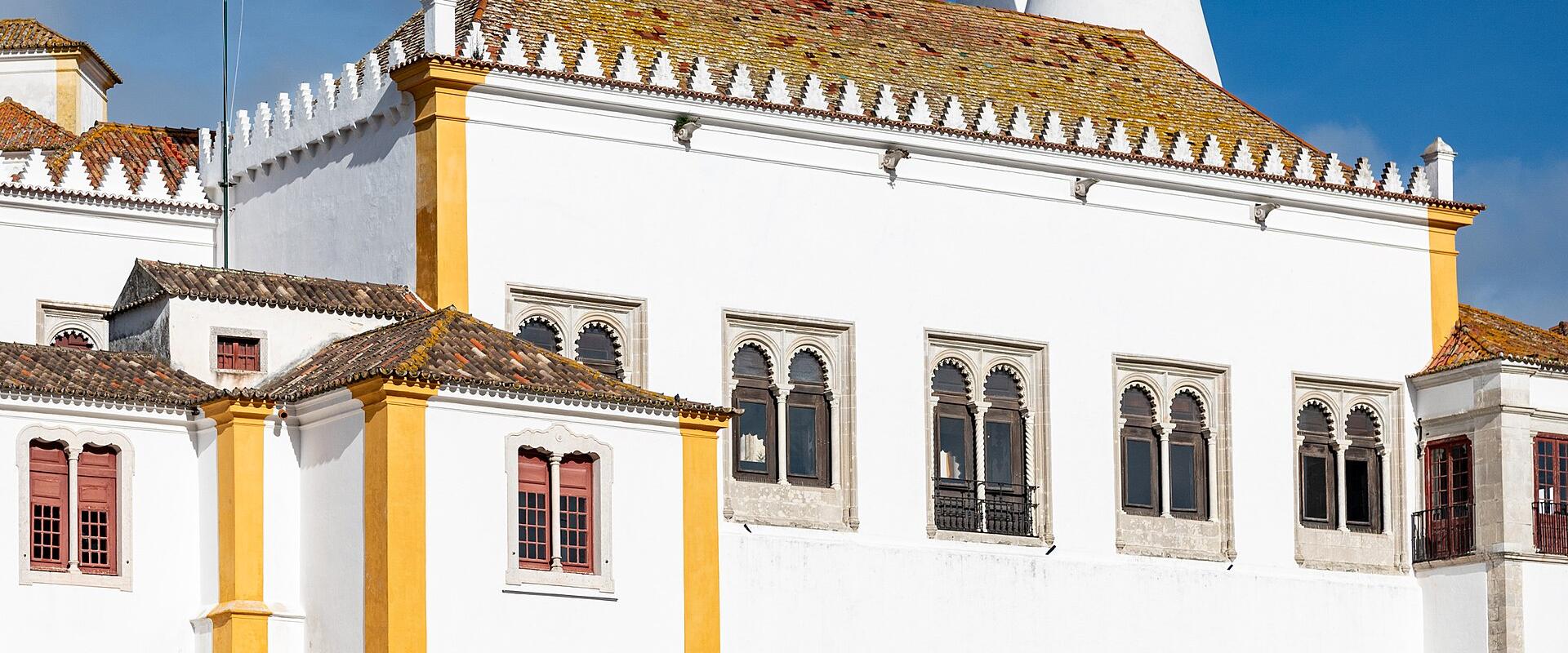 A white and yellow historic building with Gothic arches, a crenellated roofline, two large white conical chimneys, and a Portuguese flag flying on a pole.