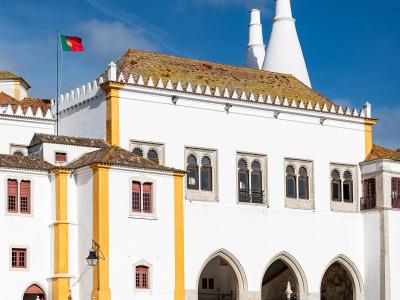 A white and yellow historic building with Gothic arches, a crenellated roofline, two large white conical chimneys, and a Portuguese flag flying on a pole.