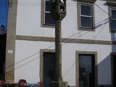 A stone pillory (pelourinho) with a multi-sided capital stands on a stepped base in a paved village square, in front of a white building with stone trim and blue doors.