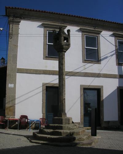 A stone pillory (pelourinho) with a multi-sided capital stands on a stepped base in a paved village square, in front of a white building with stone trim and blue doors.