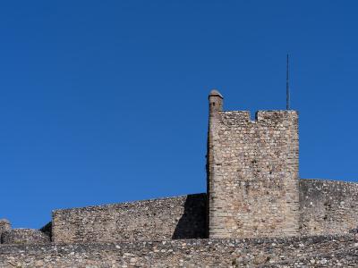 A close-up view of a medieval stone fortification featuring a square tower with crenellations and a smaller turret, set against a clear blue sky.