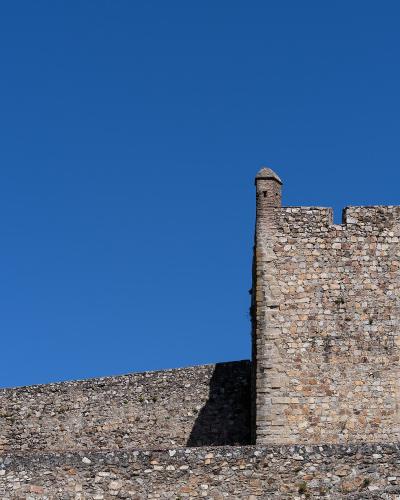 A close-up view of a medieval stone fortification featuring a square tower with crenellations and a smaller turret, set against a clear blue sky.