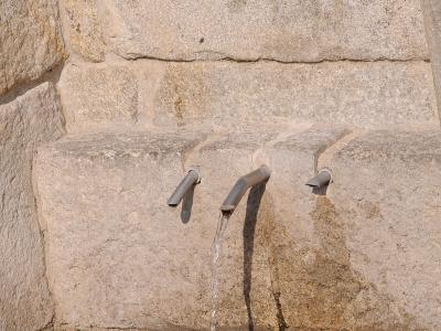 A close-up view of three metal spouts protruding from a rough-hewn stone wall, with water flowing from the central spout into a metal grate drain below.