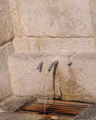 A close-up view of three metal spouts protruding from a rough-hewn stone wall, with water flowing from the central spout into a metal grate drain below.