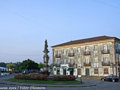 A town square featuring a central stone monument with a statue on a pedestal, surrounded by a flower bed, with a large, weathered historic building to the right and other buildings and parked cars in the background.