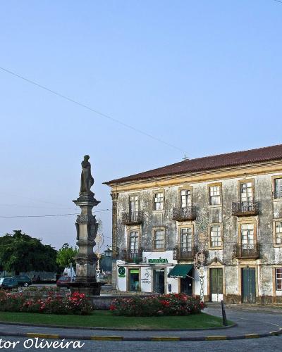 A town square featuring a central stone monument with a statue on a pedestal, surrounded by a flower bed, with a large, weathered historic building to the right and other buildings and parked cars in the background.