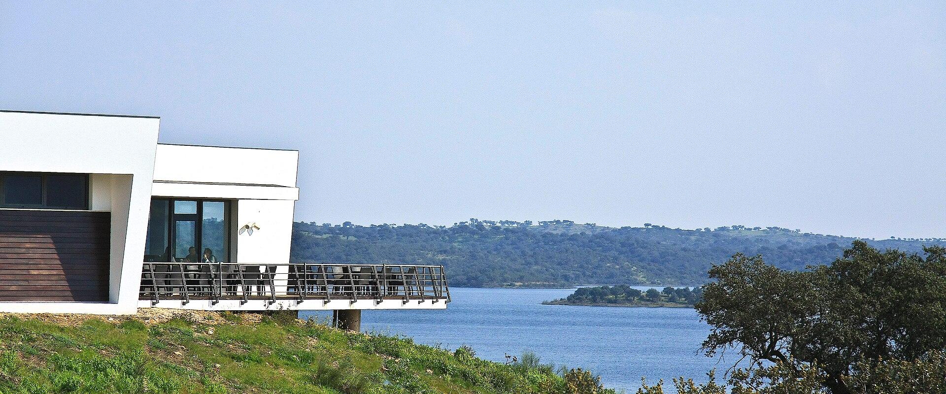 A modern white building with a cantilevered terrace and outdoor seating overlooks a wide river or estuary with a forested hillside on the opposite bank.