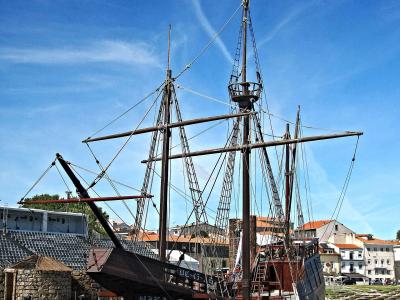 A wooden replica of a 16th-century carrack (nau) moored in a river or harbor, with a modern amphitheater and town buildings in the background.
