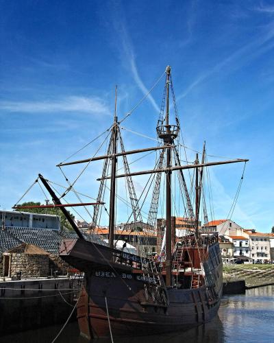 A wooden replica of a 16th-century carrack (nau) moored in a river or harbor, with a modern amphitheater and town buildings in the background.