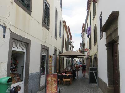 A narrow cobblestone street in a historic district lined with white buildings, featuring a man sitting on a step, outdoor restaurant seating under an umbrella, and a green trash bin.