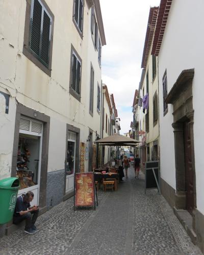 A narrow cobblestone street in a historic district lined with white buildings, featuring a man sitting on a step, outdoor restaurant seating under an umbrella, and a green trash bin.