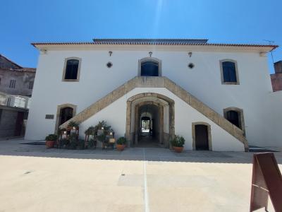 A white, two-story building with a prominent external stone staircase leading to an upper-level entrance, featuring arched windows and a central arched passageway at ground level, set against a clear blue sky.
