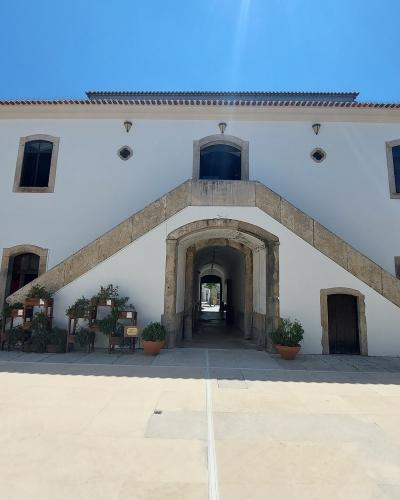 A white, two-story building with a prominent external stone staircase leading to an upper-level entrance, featuring arched windows and a central arched passageway at ground level, set against a clear blue sky.