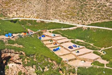 An aerial view of a campsite with tents pitched on stone-paved terraces built into a grassy, rocky hillside, with a dirt road and cliffs visible.