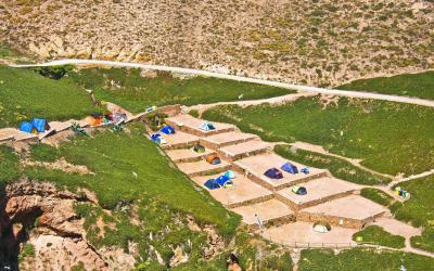 An aerial view of a campsite with tents pitched on stone-paved terraces built into a grassy, rocky hillside, with a dirt road and cliffs visible.