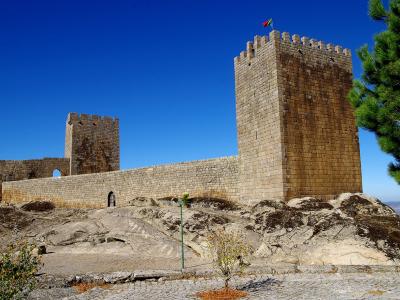 A stone medieval castle with two prominent square towers and crenellated walls situated on a rocky outcrop under a clear blue sky, with a Portuguese flag flying from the main tower.