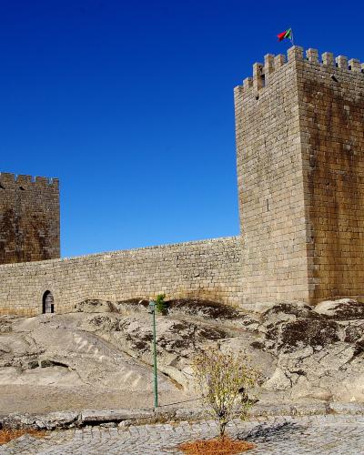 A stone medieval castle with two prominent square towers and crenellated walls situated on a rocky outcrop under a clear blue sky, with a Portuguese flag flying from the main tower.