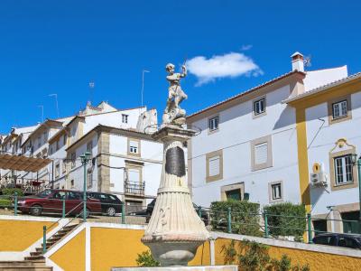 A stone fountain with a statue of a figure holding a trident (resembling Neptune or a river god) stands in a paved town square with white and yellow buildings in the background.