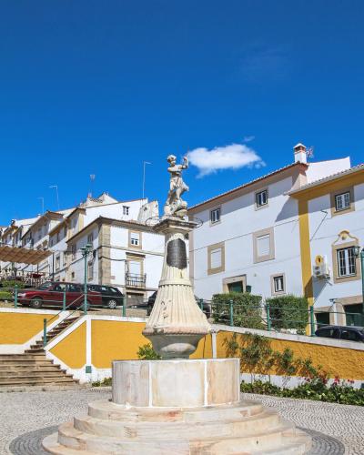 A stone fountain with a statue of a figure holding a trident (resembling Neptune or a river god) stands in a paved town square with white and yellow buildings in the background.