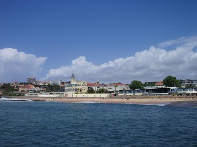 A coastal scene showing a sandy beach with gentle waves, backed by a promenade and a row of buildings including a prominent yellow structure with a pointed tower, under a blue sky with clouds.