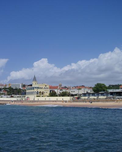 A coastal scene showing a sandy beach with gentle waves, backed by a promenade and a row of buildings including a prominent yellow structure with a pointed tower, under a blue sky with clouds.