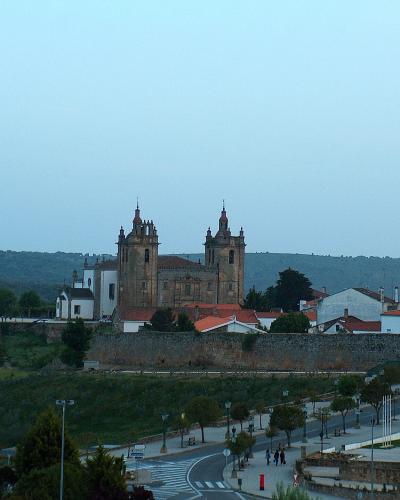 A wide shot of a large stone cathedral with two prominent towers and a fortified wall, situated on a hill overlooking a town and a riverbank promenade.