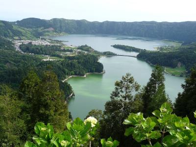 A high-angle landscape photograph showing a large volcanic caldera containing two distinct lakes separated by a narrow isthmus and a bridge, with a town visible on the left shore and lush green vegetation in the foreground.