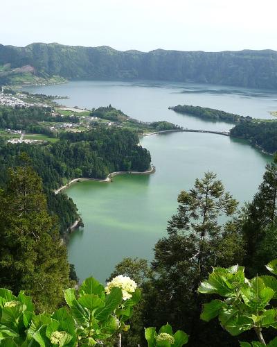 A high-angle landscape photograph showing a large volcanic caldera containing two distinct lakes separated by a narrow isthmus and a bridge, with a town visible on the left shore and lush green vegetation in the foreground.