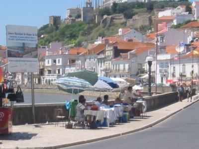 A riverside promenade with outdoor dining tables under umbrellas, a large informational sign for "Câmara Municipal de Alcácer do Sal" on the left, and a hillside town with a castle ruin in the background.