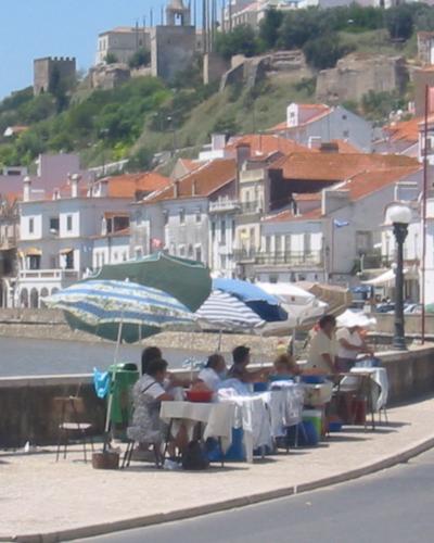 A riverside promenade with outdoor dining tables under umbrellas, a large informational sign for "Câmara Municipal de Alcácer do Sal" on the left, and a hillside town with a castle ruin in the background.
