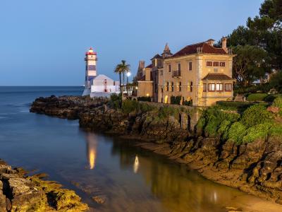 A coastal scene at twilight featuring the Santa Marta Lighthouse with its distinctive blue and white horizontal stripes, situated next to a large historic building (the Casa do Guindaste) on a rocky promontory overlooking the ocean.