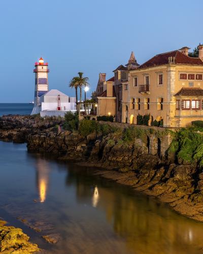 A coastal scene at twilight featuring the Santa Marta Lighthouse with its distinctive blue and white horizontal stripes, situated next to a large historic building (the Casa do Guindaste) on a rocky promontory overlooking the ocean.