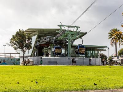 A cable car station with two blue gondolas at the lower terminal, featuring a green metal support structure, a glass-walled waiting area, and a grassy foreground with people and pigeons.