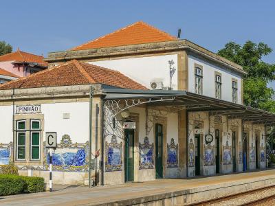 A white railway station building with a terracotta tiled roof, decorated with traditional blue and white azulejo tiles, featuring a platform sign that reads "PINHÃO".