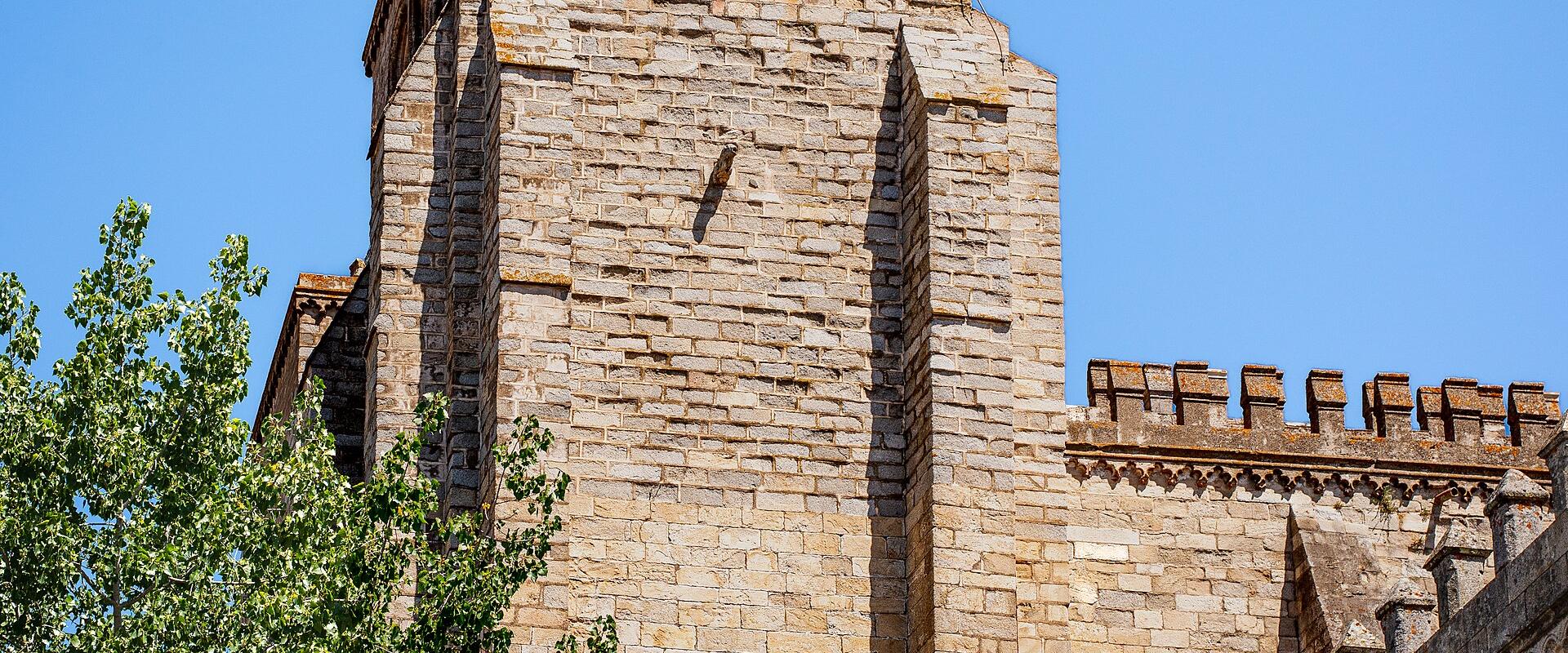 A low-angle view of a medieval stone bell tower with two visible bells in arched openings, crenellated parapets, and a green tree in the foreground against a clear blue sky.