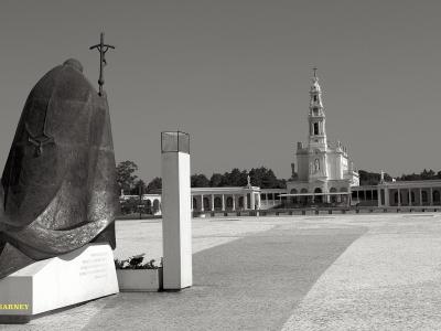 A black and white photograph showing a large bronze statue of the Virgin Mary in the foreground and the Basilica of Our Lady of the Rosary in the background, situated within a large paved plaza.