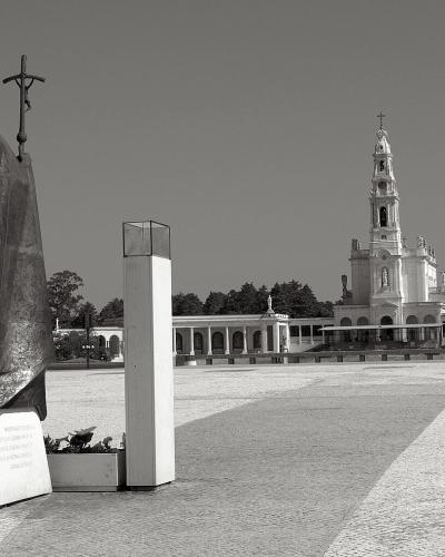 A black and white photograph showing a large bronze statue of the Virgin Mary in the foreground and the Basilica of Our Lady of the Rosary in the background, situated within a large paved plaza.