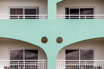 A close-up architectural detail of a hotel building featuring mint-green structural arches, white railings, and sliding glass doors with dark curtains.