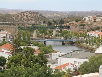 A high-angle view of a river valley in a Portuguese town featuring three distinct bridges: a stone aqueduct with a green metal truss section, a lower concrete road bridge, and a modern highway viaduct in the background.
